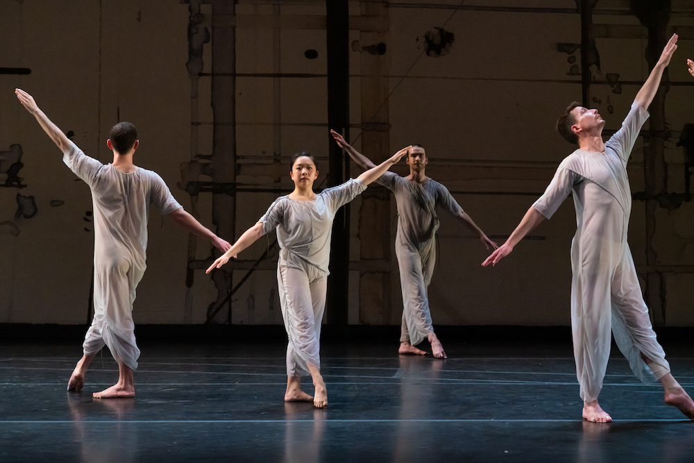 four dancers stand on a stage wearing sheer blue-grey jump suits and pointing one foot forward, .they look directly into the audience with their arms spread in diagonal line from fingertip to finger tip...another dancer stands facing a side corner. we see him in profile standing similarly to the other three, his gaze follows the path if his higher arm, his lower arm behind him finishes the diagonal line